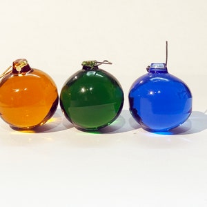 Op de afbeelding: Drie glazen kerstballen in verschillende kleuren: amber, groen en blauw. Elke bal heeft een metalen hanger.