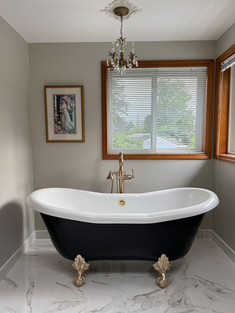 May include: A black clawfoot bathtub with gold accents sits on a white marble floor in a bathroom with a white ceiling and gray walls. A crystal chandelier hangs from the ceiling.