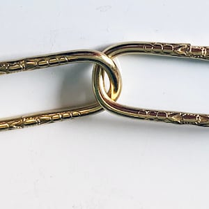 May include: Close-up of a gold chain with oval links. The links have a decorative pattern.