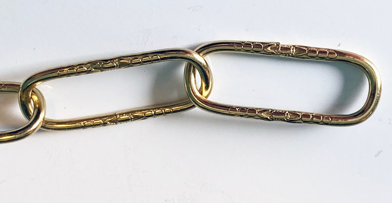 May include: Close-up of a gold chain with oval links. The links have a decorative pattern.