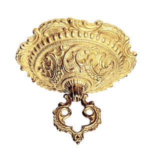Puede incluir: Un florón de techo de metal dorado con un patrón ornamentado y en relieve de flores y volutas. La placa circular presenta un borde festoneado y una abertura central roscada, de la que cuelga un anillo decorativo de metal dorado a juego.