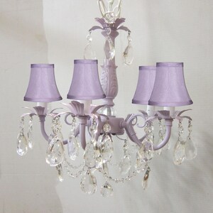 May include: A lavender chandelier with five light bulbs and crystal accents. The chandelier has a traditional design with a central stem and five arms that extend outwards. Each arm has a lavender fabric lampshade and a crystal pendant.