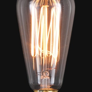Op de afbeelding: Een vintage stijl gloeilamp met een heldere glazen behuizing en een warm gloeiende gloeidraad. De lamp heeft een messing voet en een druppelvorm, ideaal voor decoratieve verlichting. De gloeidraad is een reeks verticale en horizontale lijnen.