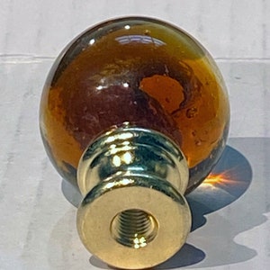 May include: A round amber glass finial with a gold metal base. The finial has a threaded bottom for attaching to a curtain rod.