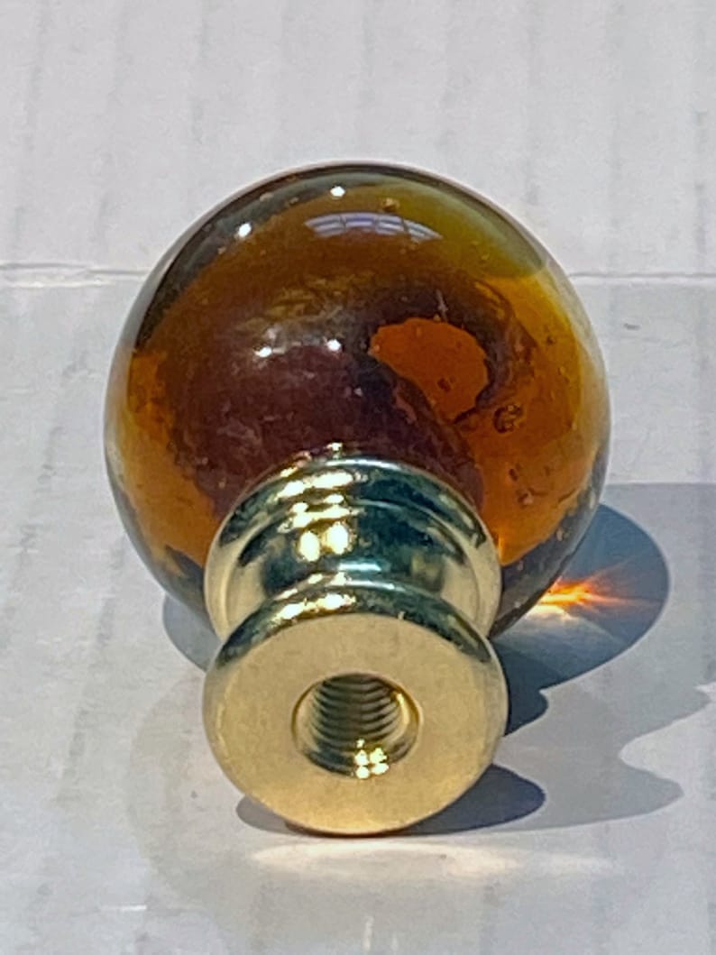 May include: A round amber glass finial with a gold metal base. The finial has a threaded bottom for attaching to a curtain rod.