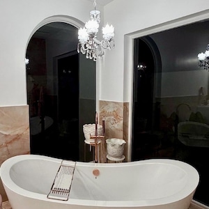 May include: A white freestanding oval bathtub with a copper-toned faucet and a matching metal tray resting across the rim. Above the tub hangs a crystal chandelier with multiple lights, set against a backdrop of white walls and marble tiles.