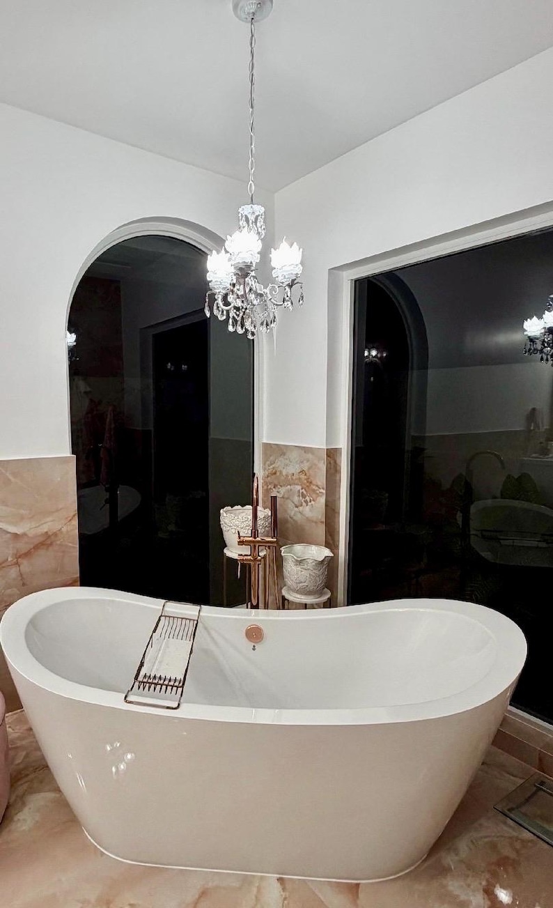 May include: A white freestanding oval bathtub with a copper-toned faucet and a matching metal tray resting across the rim. Above the tub hangs a crystal chandelier with multiple lights, set against a backdrop of white walls and marble tiles.