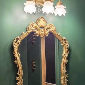 May include: A wall-mounted mirror with an ornate gold-colored frame featuring intricate scrollwork and a decorative crest at the top. Above the mirror is a three-light fixture with gold-colored metal arms and white glass shades shaped like flowers.