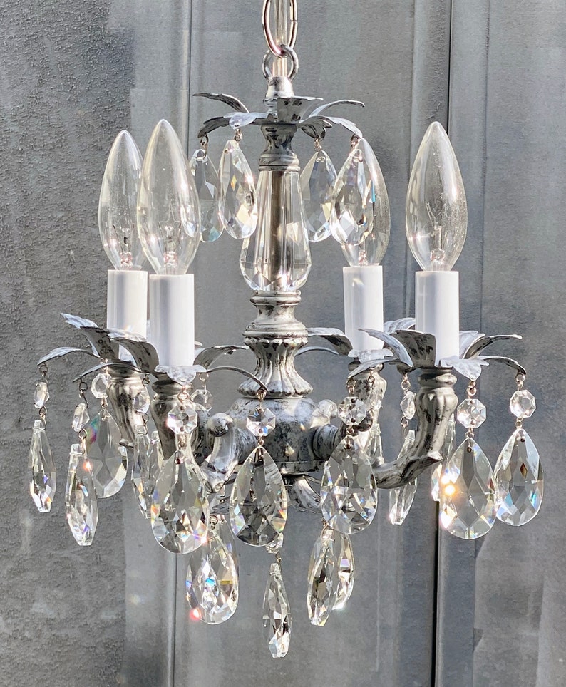 May include: A silver chandelier with crystal drops and four candle-style light bulbs. The chandelier has a distressed, aged look.