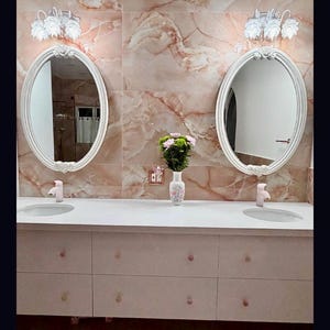 May include: A double vanity bathroom set featuring two oval mirrors with white ornate frames, two white sinks with light pink faucets, and a white cabinet base with eight drawers. A white vase with pink and green flowers sits in the center.