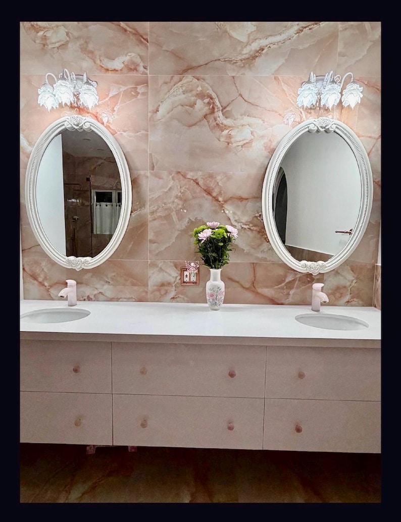May include: A double vanity bathroom set featuring two oval mirrors with white ornate frames, two white sinks with light pink faucets, and a white cabinet base with eight drawers. A white vase with pink and green flowers sits in the center.