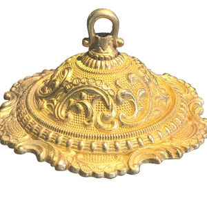 May include: A gold-coloured metal ceiling canopy with a dome shape and a central loop at the top. The surface features intricate embossed scrollwork, floral patterns, and a textured stippled background with a scalloped, decorative edge.