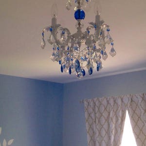 May include: A white chandelier with blue crystal accents hangs from a white ceiling. The chandelier has three light bulbs and a blue glass orb in the center.