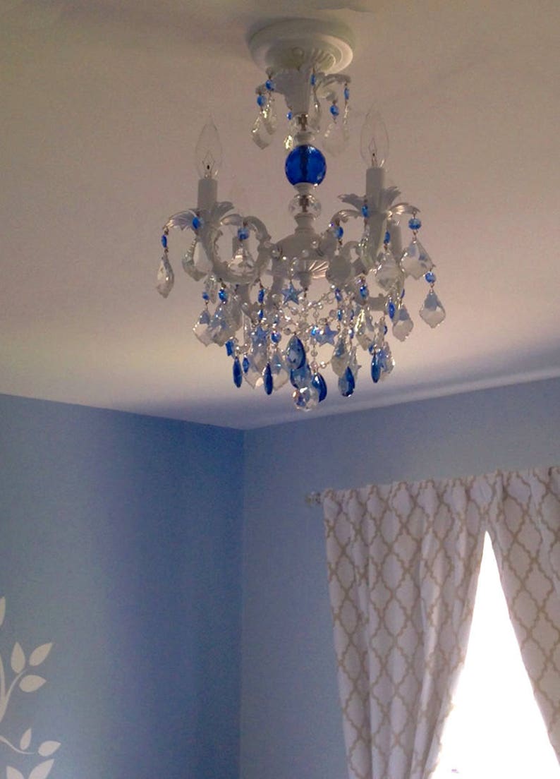 May include: A white chandelier with blue crystal accents hangs from a white ceiling. The chandelier has three light bulbs and a blue glass orb in the center.