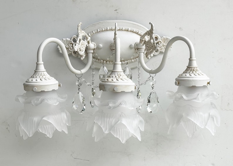 May include: A three-light wall sconce featuring a matte white finish, an oval backplate with ornate detailing, curved arms, and three frosted glass shades shaped like flower petals, accented with hanging clear crystal teardrop prisms.