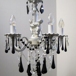 May include: A white chandelier with black crystal drops and cherub accents. The chandelier has five light bulbs and is decorated with clear and white beads.