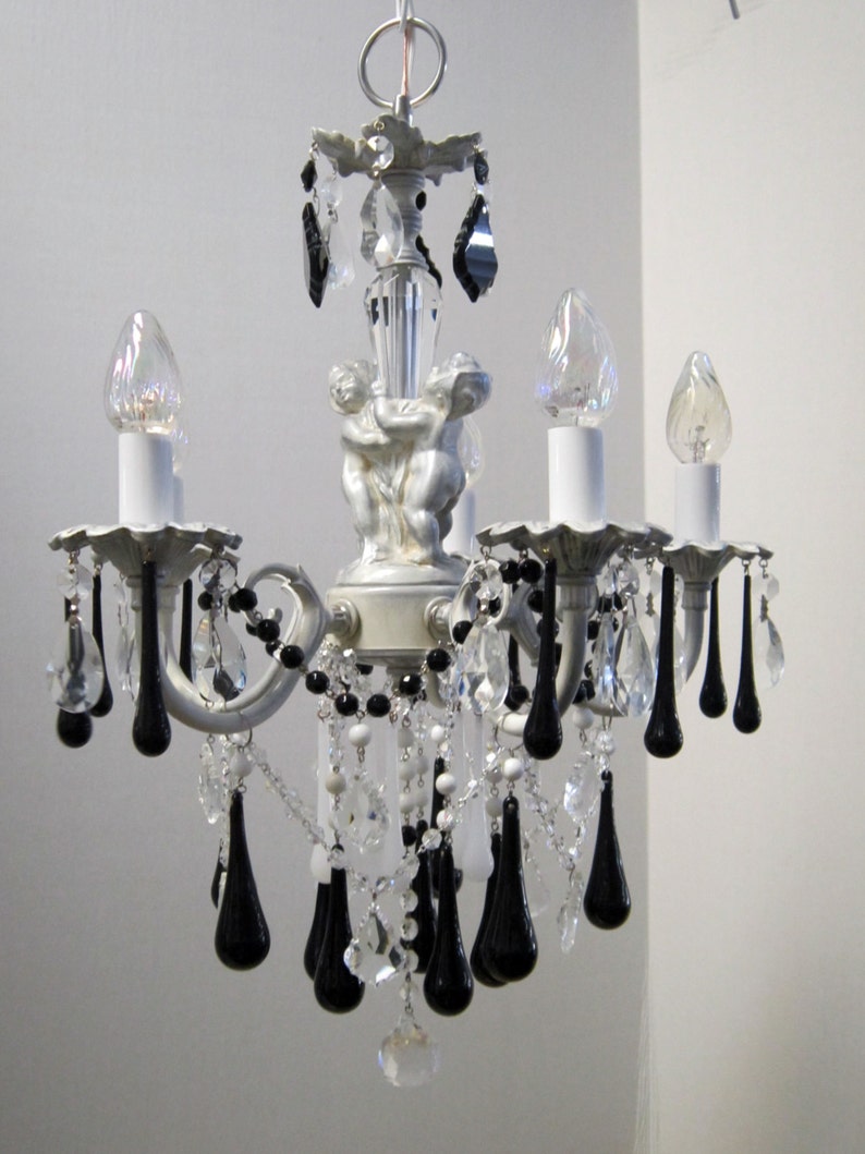 May include: A white chandelier with black crystal drops and cherub accents. The chandelier has five light bulbs and is decorated with clear and white beads.