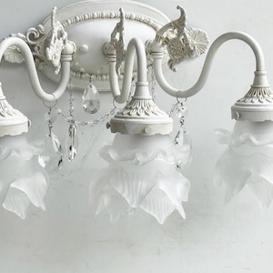 May include: A white three-light wall sconce featuring curved metal arms and an oval backplate with decorative molding. Each light is encased in a frosted glass shade shaped like a flower petal, accented by hanging clear crystal teardrop prisms.