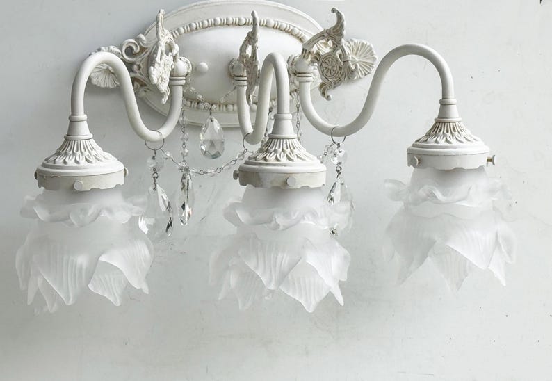 May include: A white three-light wall sconce featuring curved metal arms and an oval backplate with decorative molding. Each light is encased in a frosted glass shade shaped like a flower petal, accented by hanging clear crystal teardrop prisms.