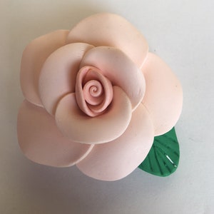 Op de afbeelding: Een roze keramische roos met een groen blad. De roos is gemaakt van meerdere lagen bloemblaadjes en heeft een spiraalvormig midden.