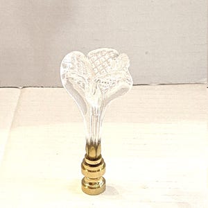 May include: A clear glass bottle stopper with a flower-shaped top. The stopper has a gold-colored metal base with a series of stacked rings. The flower design has a textured surface.