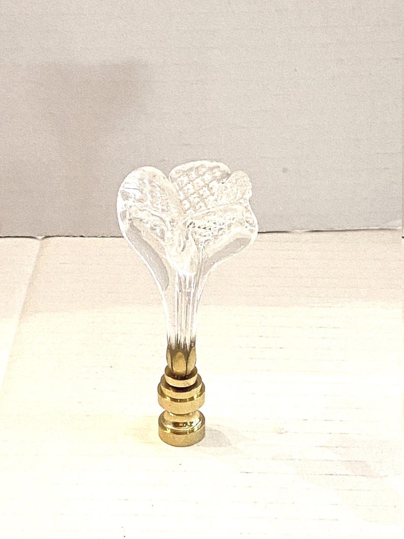 May include: A clear glass bottle stopper with a flower-shaped top. The stopper has a gold-colored metal base with a series of stacked rings. The flower design has a textured surface.