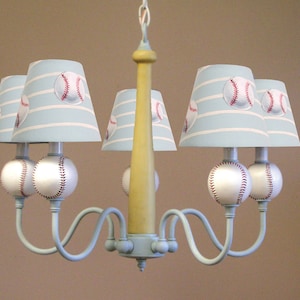 May include: A five-light chandelier with a baseball theme. The chandelier has a yellow wooden base and blue metal arms. The lampshades are light blue with white stripes and a baseball design. The light bulbs are covered with white baseballs.