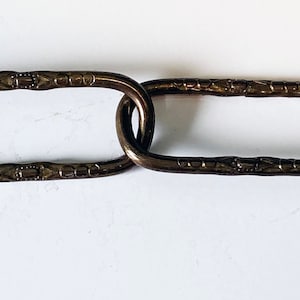 May include: Close-up of a decorative chain with oval links. The chain is made of metal and has a dark brown finish. The links have a textured design.