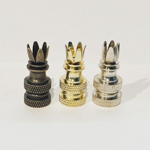 May include: Three metal lamp finials in bronze, gold, and silver. Each finial has a crown-like design at the top and a textured base. The finials are displayed against a white background.