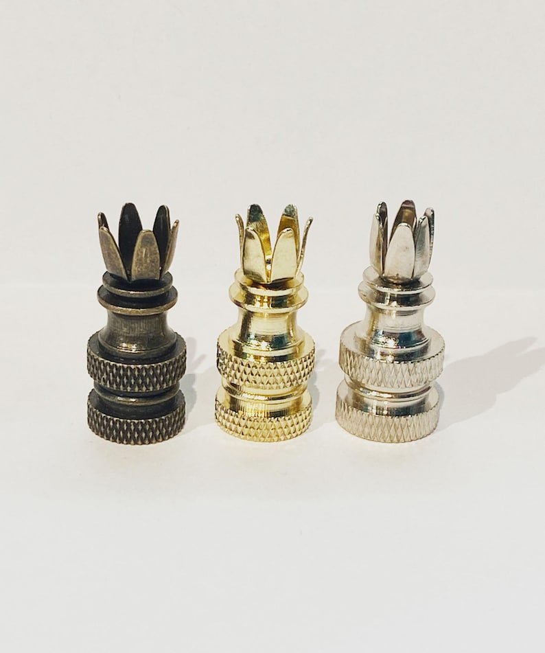 May include: Three metal lamp finials in bronze, gold, and silver. Each finial has a crown-like design at the top and a textured base. The finials are displayed against a white background.