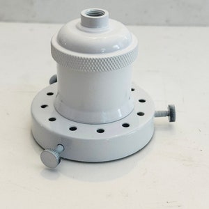 Könnte beinhalten: Eine weiße Metall-Leuchte mit einem runden Sockel und einem Gewinde oben. Der Sockel hat eine Reihe von Löchern am Rand und zwei Schrauben an der Seite.