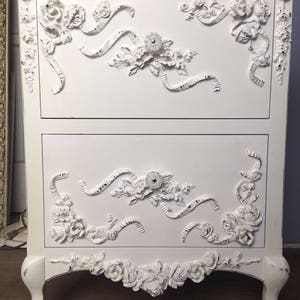 May include: White painted wooden chest of drawers with ornate floral and ribbon carvings. The chest has two drawers and sits on four legs.