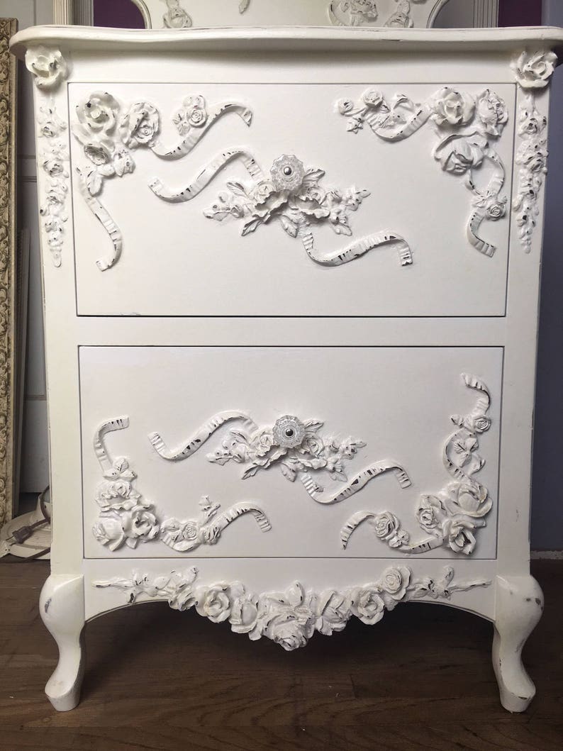 May include: White painted wooden chest of drawers with ornate floral and ribbon carvings. The chest has two drawers and sits on four legs.