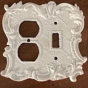 May include: White decorative outlet cover with an ornate design. The cover has two outlets and one switch.