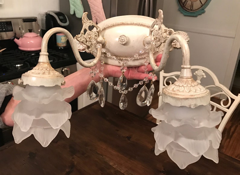 May include: A two-light wall sconce featuring an off-white, distressed metal base with ornate scrollwork. Two frosted glass shades shaped like layered flower petals hang from curved arms, accented by clear crystal teardrop drapes in the center.