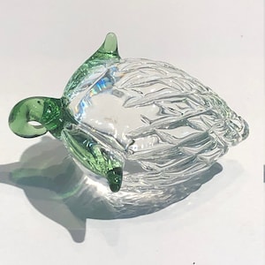 Op de afbeelding: Een heldere glazen ornament in de vorm van een vogel met groene glazen accenten. De ornament heeft een getextureerd oppervlak en een kleine lus aan de bovenkant om op te hangen.