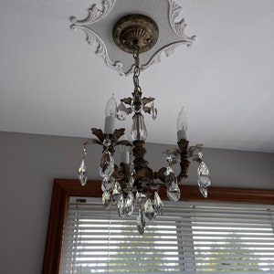 May include: A gold chandelier with crystal drops hangs from a decorative ceiling medallion. The chandelier has four candle-style light bulbs and is surrounded by a white ceiling.