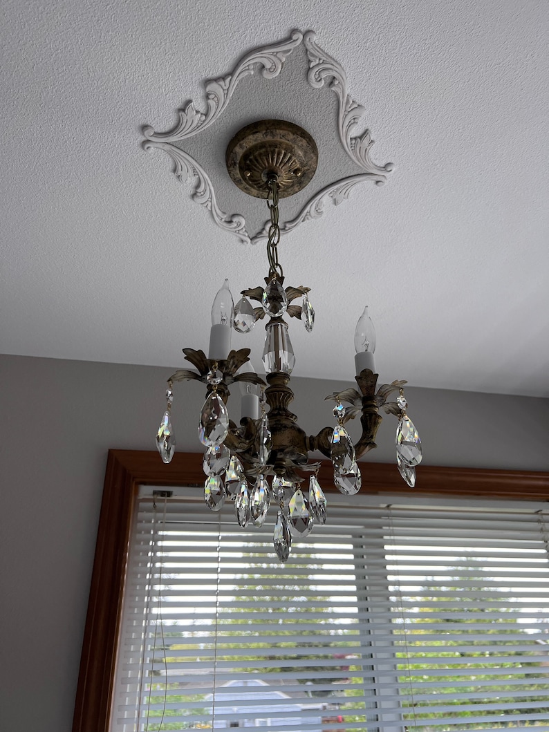 May include: A gold chandelier with crystal drops hangs from a decorative ceiling medallion. The chandelier has four candle-style light bulbs and is surrounded by a white ceiling.