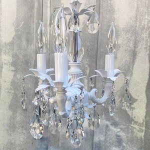 May include: White chandelier with crystal drops and candle-style light bulbs. The chandelier has a floral design and is made of metal.
