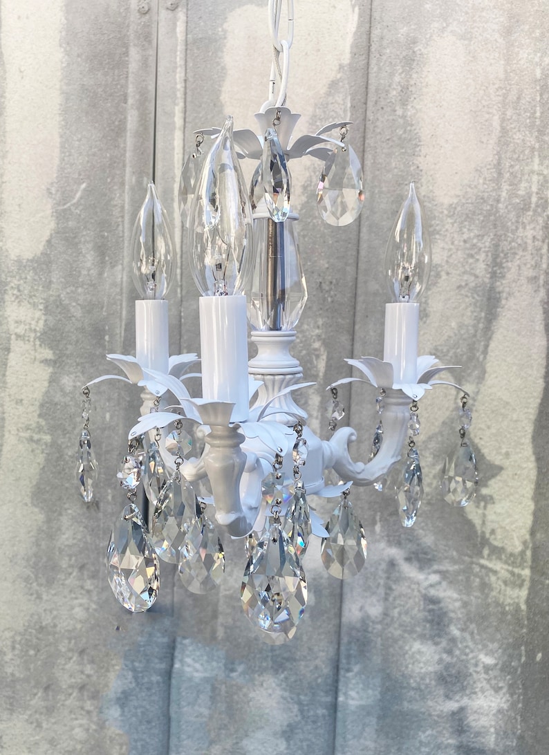 May include: White chandelier with crystal drops and candle-style light bulbs. The chandelier has a floral design and is made of metal.