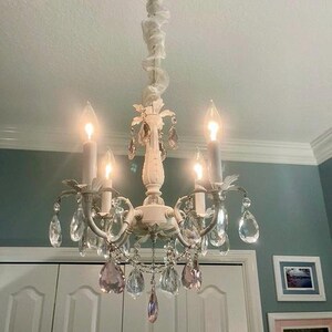 May include: A white chandelier with crystal drops and a white fabric swag hanging from the ceiling. The chandelier has four light bulbs and the crystals are clear with a hint of pink.