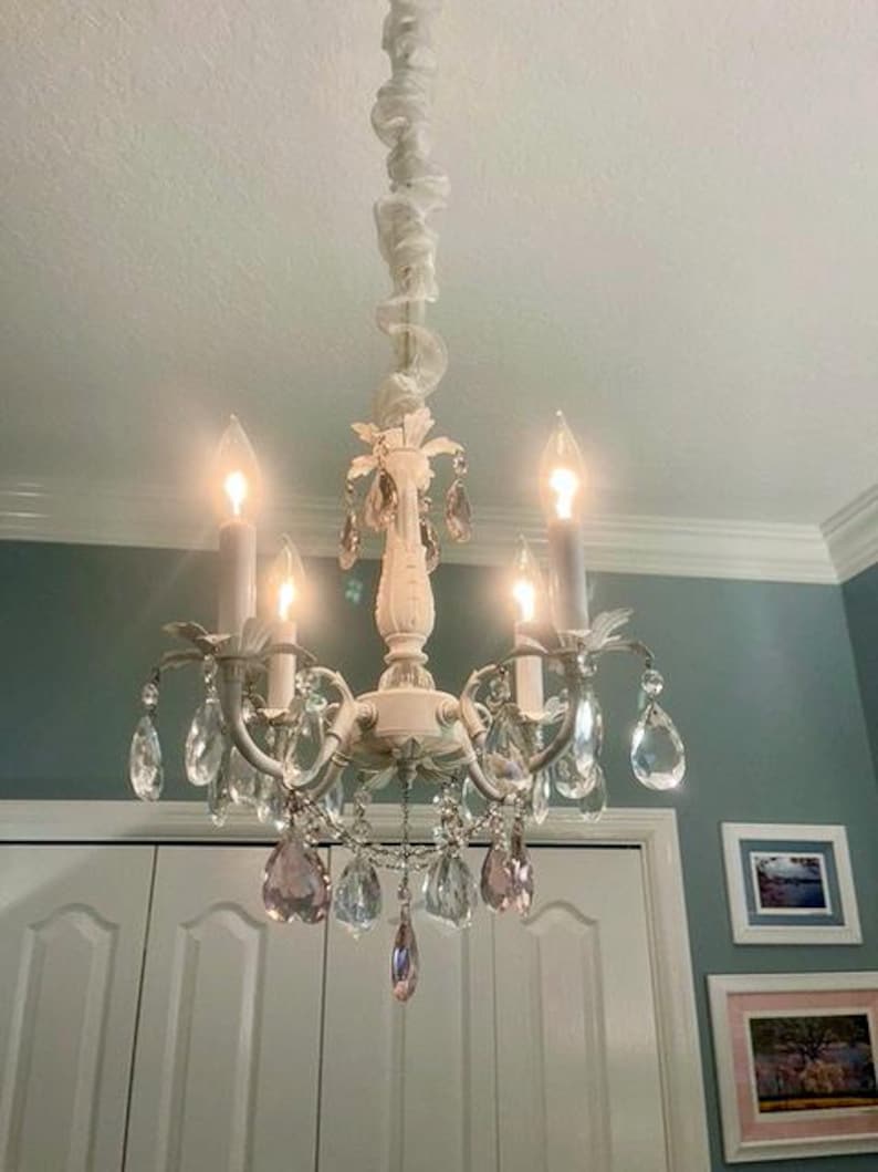May include: A white chandelier with crystal drops and a white fabric swag hanging from the ceiling. The chandelier has four light bulbs and the crystals are clear with a hint of pink.