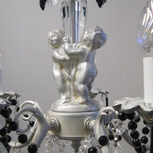 May include: A white and silver chandelier with black beads and crystal drops. The chandelier features two cherub figurines holding a crystal rod.
