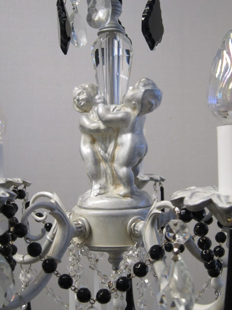 May include: A white and silver chandelier with black beads and crystal drops. The chandelier features two cherub figurines holding a crystal rod.