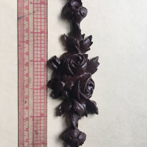 May include: A dark brown, ornate, wooden rose design with leaves. The design is approximately 7 inches tall.