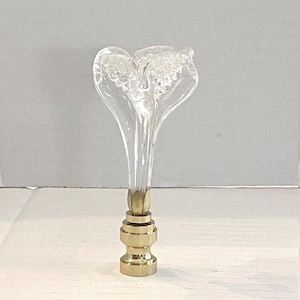May include: A clear glass lamp finial with a flower-like design. The finial has a brass base and a clear, elongated stem. The top of the finial has a textured, frosted appearance. The lamp finial is a decorative lighting accessory.