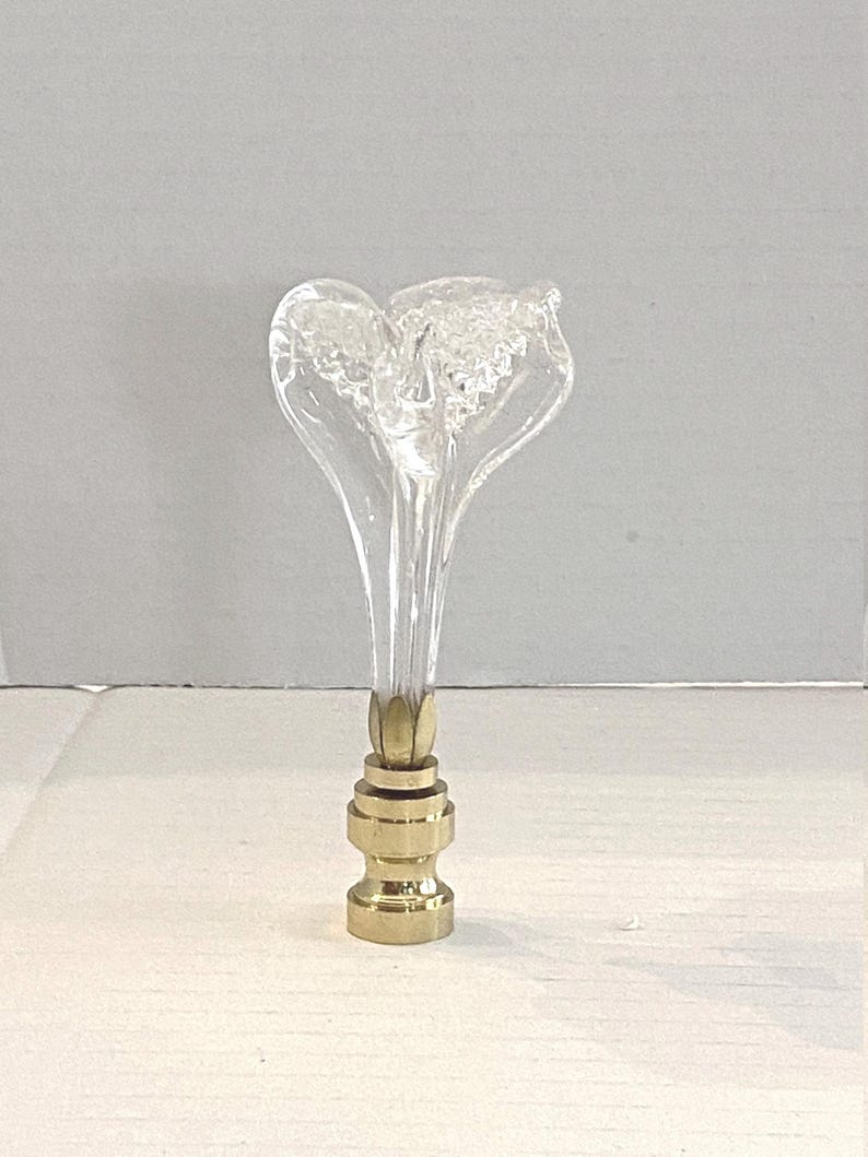 May include: A clear glass lamp finial with a flower-like design. The finial has a brass base and a clear, elongated stem. The top of the finial has a textured, frosted appearance. The lamp finial is a decorative lighting accessory.
