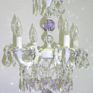 May include: White chandelier with crystal and purple glass accents. The chandelier has four candle-style light bulbs and a decorative top.