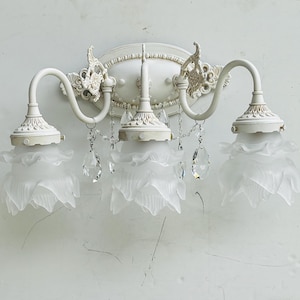 May include: A three-light wall sconce featuring a distressed off-white metal frame with ornate scrollwork and an oval backplate. Three frosted glass shades shaped like flower petals hang from curved arms, accented by clear crystal teardrop prisms.