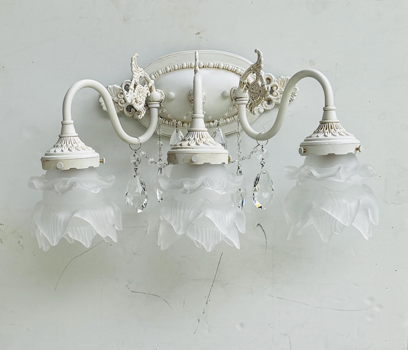 May include: A three-light wall sconce featuring a distressed off-white metal frame with ornate scrollwork and an oval backplate. Three frosted glass shades shaped like flower petals hang from curved arms, accented by clear crystal teardrop prisms.